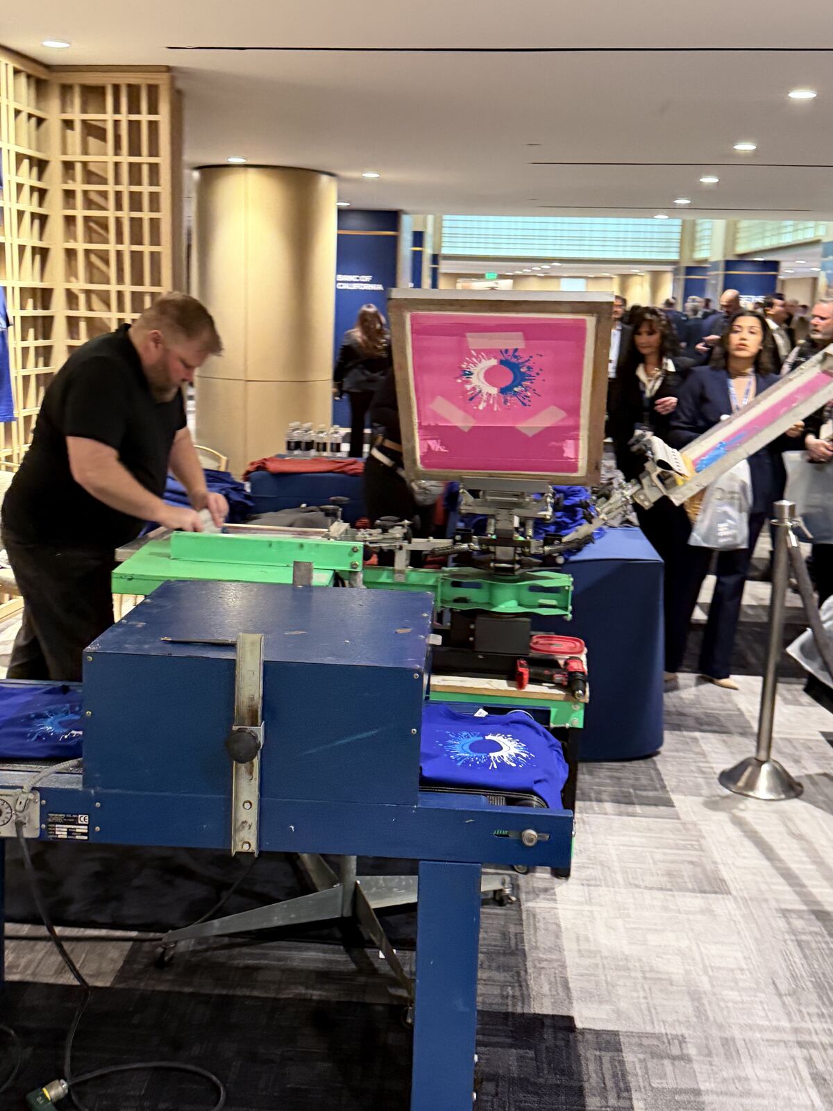 Live event demonstration showing an operator working at a screen printing and heat press station with spectators watching and display samples visible